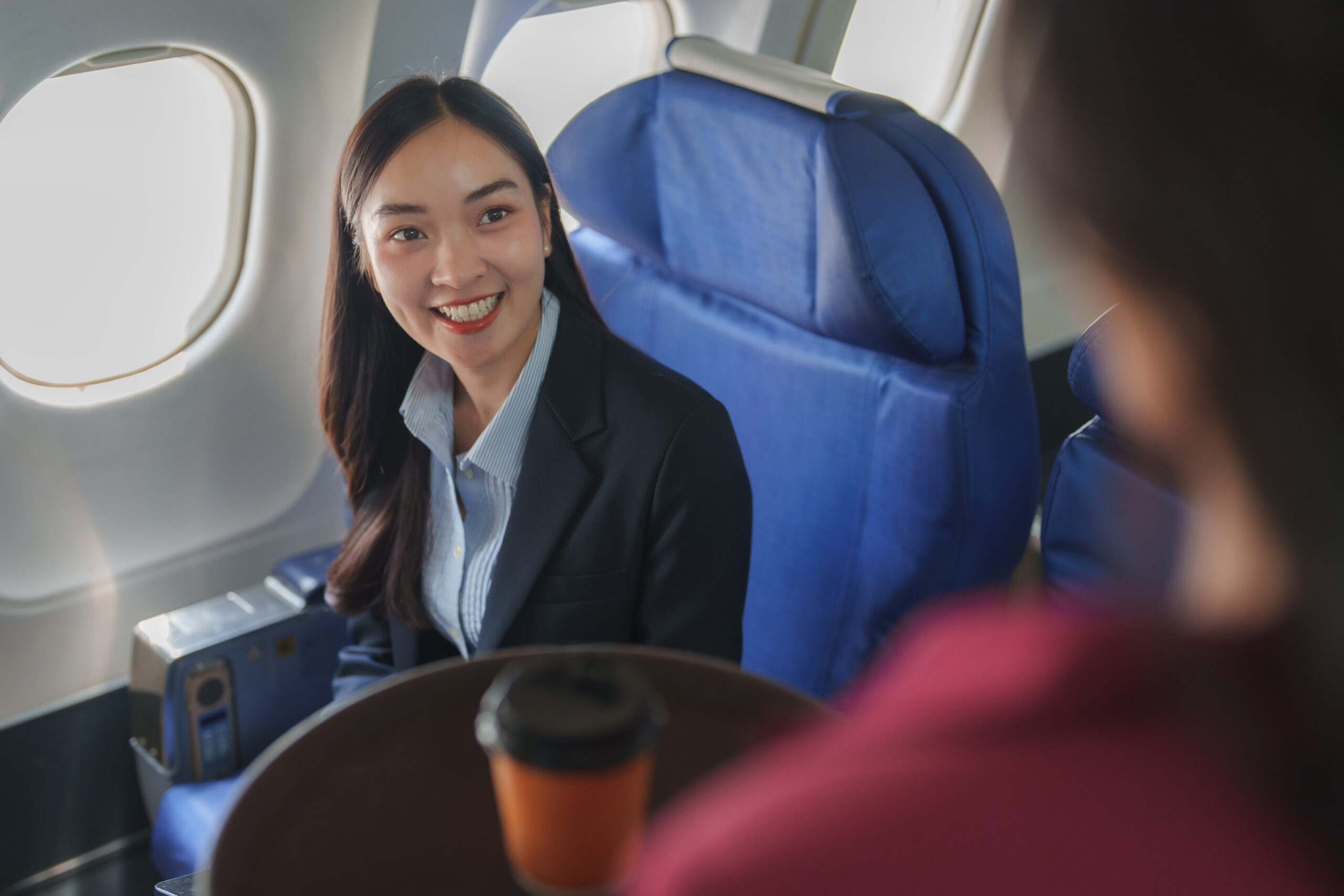 Businesswoman wearing a suit and sitting in her seat, smiling and talking to a flight attendant while enjoying a hot beverage during a comfortable flight